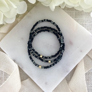 May include: A set of three beaded bracelets featuring a repeating pattern of matte black and translucent dark gray seed beads. Each bracelet is accented with a single polished gold-tone metal bead positioned at the center of the strand.