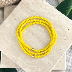 May include: A triple-strand bracelet composed of small, vibrant yellow seed beads. The design features three distinct circular rows of beads, each accented with a single, polished gold-toned metallic sphere bead integrated into the strand.