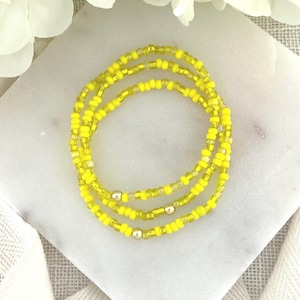 May include: A multi-strand bracelet composed of small, vibrant yellow seed beads. The design features several smooth, metallic gold-toned spherical beads interspersed throughout the yellow bead strands, creating a bright and uniform circular accessory.