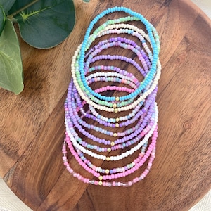 May include: A collection of beaded bracelets in various pastel colors, including pink, purple, blue, and white. The bracelets are made of small, round beads and are arranged on a wooden surface. Each bracelet has a gold-colored clasp.