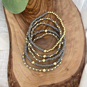 May include: Stack of beaded bracelets. The bracelets feature gray beads and gold-colored accent beads. The bracelets are displayed on a wooden surface.