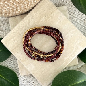 May include: A multi-strand beaded bracelet featuring a collection of small, round glass beads in shades of deep burgundy, dark maroon, and metallic gold. The strands are arranged in a circular formation, showcasing a mix of matte and shiny finishes.