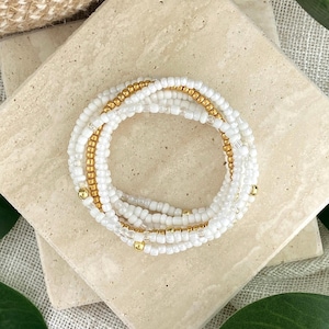 May include: A circular bracelet composed of multiple strands of small white seed beads interspersed with metallic gold beads. The design features a repeating pattern of white and gold, creating a layered appearance with a polished finish.