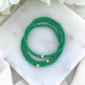 May include: A set of three stretch bracelets made of small, round, vibrant green beads. Each bracelet features a single, smooth, polished gold-tone metal bead integrated into the strand of green beads, creating a consistent and uniform appearance.