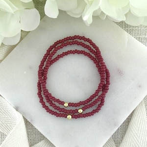 May include: Three burgundy beaded bracelets with gold accents are arranged on a white marble surface. The bracelets are made of small, round beads and are wrapped around each other. The gold accents are small, round beads that are spaced evenly on each bracelet.