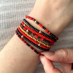 May include: A stack of multiple beaded bracelets featuring small round beads in alternating patterns of solid black, vibrant red, and metallic gold. The bracelets are arranged in a layered design, showcasing a consistent color scheme throughout.