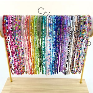 May include: A display of colorful beaded necklaces in a variety of colors, including red, pink, orange, yellow, green, blue, purple, and brown. The necklaces are hanging from a gold-colored stand with a wooden base. The beads are small and uniform in size.