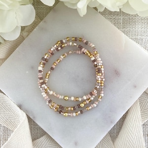 May include: Three beaded bracelets in shades of pink, white, brown, and gold. The bracelets are made of small, round beads strung together. The bracelets are stacked on a white marble surface, with a white flower and ribbon in the background.