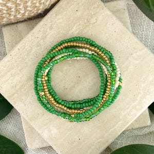 May include: A stack of four beaded bracelets featuring a mix of small round beads in shades of bright green, translucent light green, metallic gold, and opaque cream, arranged in a circular pattern on a square stone surface.
