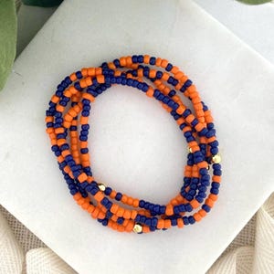 May include: A collection of orange and navy blue beaded bracelets. The bracelets are made of small, round beads and are strung together. Gold-colored beads are interspersed throughout the design. The bracelets are arranged in a circular pattern.