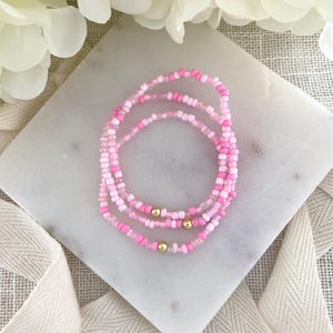 May include: Three beaded bracelets in shades of pink and white with gold-colored accents. The bracelets are arranged in a circular pattern on a white marble surface, with a soft, neutral background.