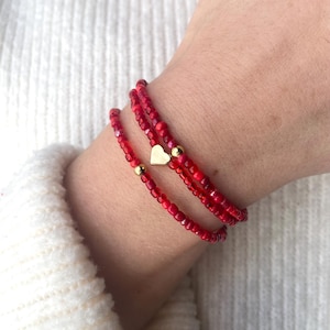 May include: Three red beaded bracelets with a gold heart charm and gold beads. The bracelets are wrapped around a wrist. The beads are small and the color is a vibrant red. The bracelets are worn over a white sweater.