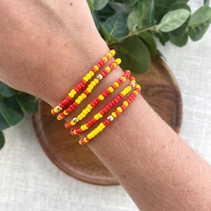 May include: A set of five beaded bracelets featuring alternating red and yellow seed beads. Each bracelet is accented with a single small, round, metallic gold-toned bead positioned at intervals along the strand of colorful glass beads.