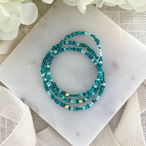 May include: A multi-strand bracelet composed of small glass seed beads in various shades of teal, turquoise, and light blue. The design features three small, smooth, metallic gold-toned spherical beads spaced evenly along the circular beaded strands.
