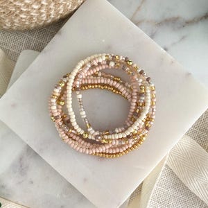 May include: A stack of multiple beaded bracelets featuring a color palette of cream, soft pink, and metallic gold. The bracelets are composed of small, uniform round beads interspersed with shiny gold-toned spherical accents throughout the strands.