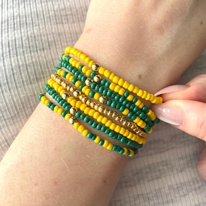 May include: A stack of seven beaded bracelets featuring alternating rows of vibrant yellow and deep forest green seed beads, accented with small, polished gold-toned metallic spheres interspersed throughout the design.