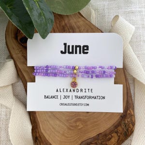 May include: A stack of three beaded bracelets in shades of purple, with a gold charm and a pink stone. The bracelets are displayed on a wooden surface with a card that reads "June" and "Alexandrite". The card also includes the words "Balance | Joy | Transformation".