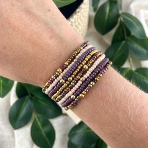 May include: A stack of seven beaded bracelets featuring a mix of small round beads in shades of purple, mauve, cream, and metallic gold. The bracelets are arranged in a layered pattern, alternating between solid matte colors and shiny metallic gold accents.