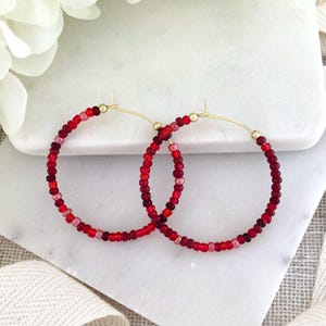 May include: A pair of hoop earrings with gold-colored posts. The hoops are adorned with small, colorful beads in shades of red and pink. The earrings are displayed on a white marble surface.
