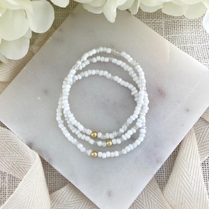 May include: Three white beaded bracelets with gold-colored accents are arranged on a white marble surface. The bracelets are made of small, round white beads. The gold accents are small, round beads. The background includes white flowers and a white ribbon.