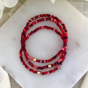May include: A triple-strand bracelet composed of small, round glass beads in various shades of red, ranging from light pinkish-red to deep crimson. Three smooth, polished gold-toned spherical metal beads are evenly spaced throughout the strands.