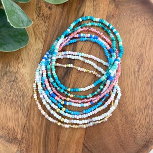 May include: A collection of beaded bracelets in various colors, including turquoise, pink, white, and gold. The bracelets are arranged in a circular pattern on a wooden surface, showcasing a mix of vibrant and pastel hues.