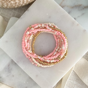 May include: A stack of circular bracelets made from small beads in shades of light pink, cream, and metallic gold. The bracelets feature a mix of opaque, translucent, and shiny finishes, arranged in a layered, circular formation.