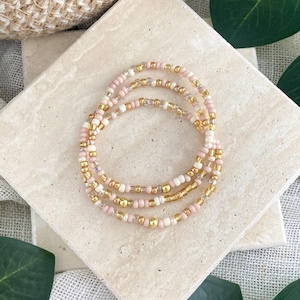 May include: A set of three beaded bracelets featuring a repeating pattern of small round beads in soft pink, cream, and metallic gold. The beads are strung on a flexible cord, creating a circular shape with a delicate and uniform appearance.