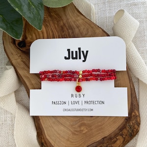 May include: A red beaded bracelet with a ruby charm, displayed on a wooden surface. The card reads "July" and "Ruby: Passion | Love | Protection". The bracelet is made of small red beads and has a gold-colored clasp.