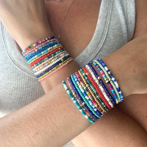 May include: Two stacks of multi-colored seed bead bracelets worn on the wrists. Each stack consists of numerous thin, elasticized strands featuring a vibrant array of opaque, translucent, and metallic gold beads in various shades of blue, red, pink, green, and white.