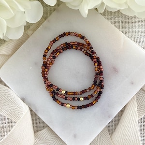 May include: A triple-strand beaded bracelet featuring small, round glass beads in shades of deep burgundy, copper, and bronze. The design is accented with three smooth, polished gold-tone metal beads spaced evenly along the strands of the bracelet.