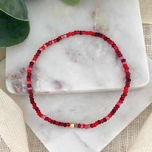 May include: A beaded bracelet featuring a gradient of red hues, from light pink to deep burgundy. The bracelet is composed of small, round beads and includes a single gold-colored bead. The jewelry is displayed on a white marble surface.
