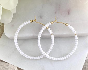 May include: A pair of hoop earrings featuring a thin gold-toned metal wire frame adorned with small, uniform white beads. The circular design is simple and consistent, with the beads covering the entire circumference of the gold metal hoop structure.
