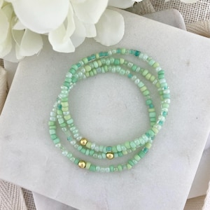 May include: Three beaded bracelets in shades of green and gold. The bracelets are made of small, round beads in various shades of green, with gold-colored beads interspersed throughout. The bracelets are arranged in a circular pattern.