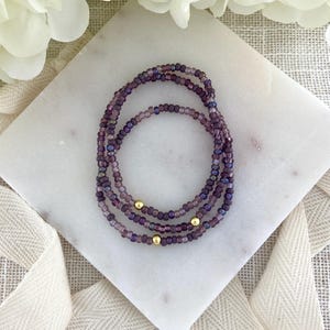 May include: Three beaded bracelets in shades of purple and gold. The bracelets are made of small, round beads and feature gold accents. The bracelets are arranged in a circular pattern on a white marble surface.
