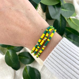 May include: A multi-strand bracelet featuring alternating yellow and green seed beads arranged in a repeating pattern, accented with small, polished gold-toned metallic beads spaced evenly throughout the design.