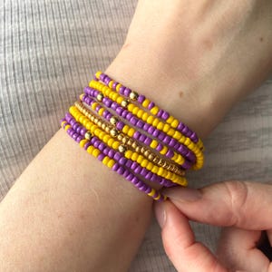 May include: A stack of multiple beaded bracelets featuring alternating rows of small purple and yellow glass beads. Several of the bracelets incorporate shiny gold-toned metallic spacer beads interspersed throughout the circular bead patterns.
