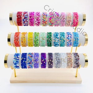 May include: A display of 24 colorful beaded bracelets with gold accents. The bracelets are arranged in three rows on a wooden stand with gold metal accents.