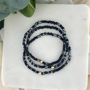 May include: Three beaded bracelets in black and gray, with gold-colored accents. The bracelets are arranged in a circular pattern on a white surface, showcasing the jewelry's design and color contrast. The beads appear to be small and uniform in size.