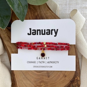 May include: A red beaded bracelet with a garnet charm, displayed on a white card that reads "January" and "Garnet." The card also lists "Courage | Faith | Authority." The bracelet is presented on a wooden surface.