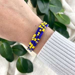 May include: A set of three stretch bracelets made from small, round, vibrant yellow and royal blue glass beads. Each bracelet features a single, polished, metallic gold-toned spherical bead integrated into the pattern of colored beads.