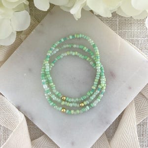 May include: Three beaded bracelets in shades of green and gold. The bracelets are made of small, round beads in various shades of green, with a few gold-colored beads interspersed. The bracelets are arranged in a circular pattern on a white marble surface.