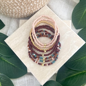May include: A stack of seven beaded bracelets featuring small round beads in shades of light pink, dark maroon, brown, and teal. Each bracelet is accented with a single small gold-toned metallic bead positioned at the center of the circular strand.