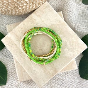 May include: A circular bracelet composed of multiple strands of small seed beads in shades of lime green, grass green, cream, and metallic gold. The beads are arranged in a layered, stacked design, creating a cohesive and colorful accessory.