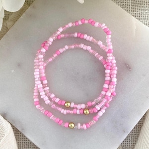 May include: A triple-strand bracelet made of small, round glass beads in various shades of pink, ranging from light pastel to vibrant magenta. The bracelet features three small, polished gold-toned metal spherical beads spaced evenly along the strands.