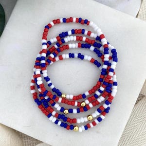May include: A collection of beaded bracelets in red, white, and blue, with gold-colored accents. The bracelets are made of small, round beads strung together. The color scheme evokes a patriotic theme.