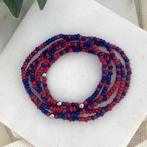 May include: Strands of red and blue beaded jewelry, possibly waist beads, are coiled on a white surface. The beads are small and round, with silver-colored metal accents. The color combination is a patriotic theme.