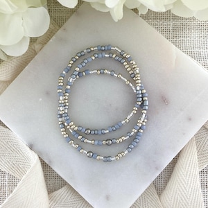 May include: Three beaded bracelets. The bracelets are made of small, light blue and silver beads. The bracelets are stacked on a white marble surface. White flowers and a white ribbon are in the background.