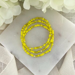 May include: Three strands of yellow beaded jewelry, possibly waist beads, are arranged in a circular pattern on a white marble surface. The beads are a bright, sunny yellow color, with small silver beads interspersed.