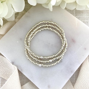 May include: A set of three circular bracelets composed of small, metallic silver-toned beads. Each bracelet features a single, slightly larger, smooth silver-toned bead positioned as a focal point among the uniform, smaller spherical beads.